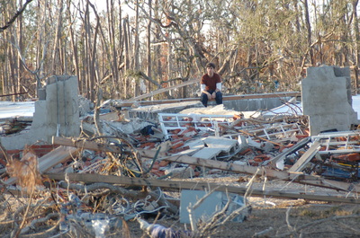 A photo of a young adult male individual sitting atop a large pile of rubble that was once a house before Katrina decimated large parts of New Orleans